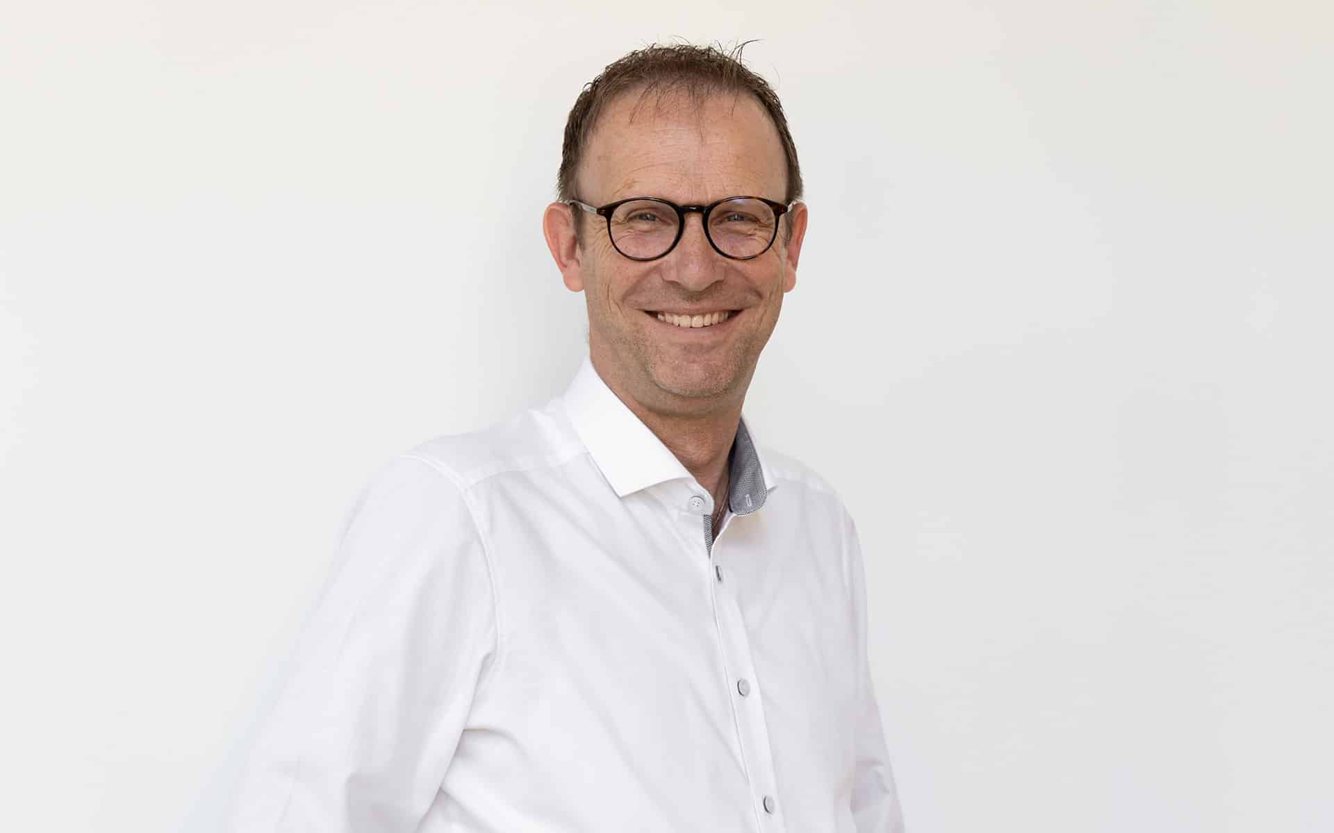 Außendienstmitarbeiter Stefan Wastl steht mit Brille und weißem Hemd vor weißer Wand und lacht.