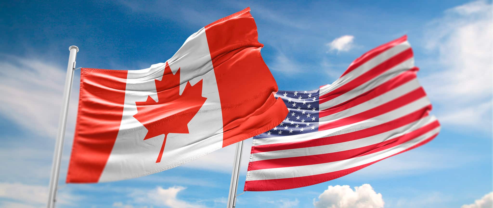 Flags of Canada and the United States in front of a blue sky with clouds.